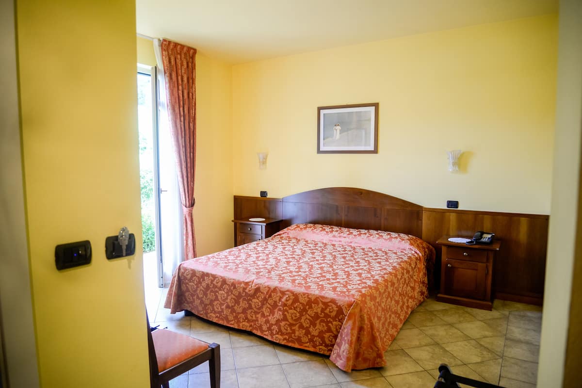 A hotel room with a bed, a wooden nightstand, a bedside table, a chair, a window with curtains, and a framed picture on the wall.
