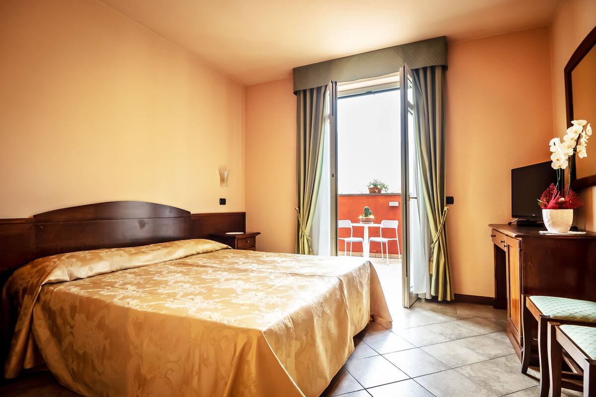 A hotel room with a large bed, a desk with a chair, a television, and a balcony with a table and chairs.