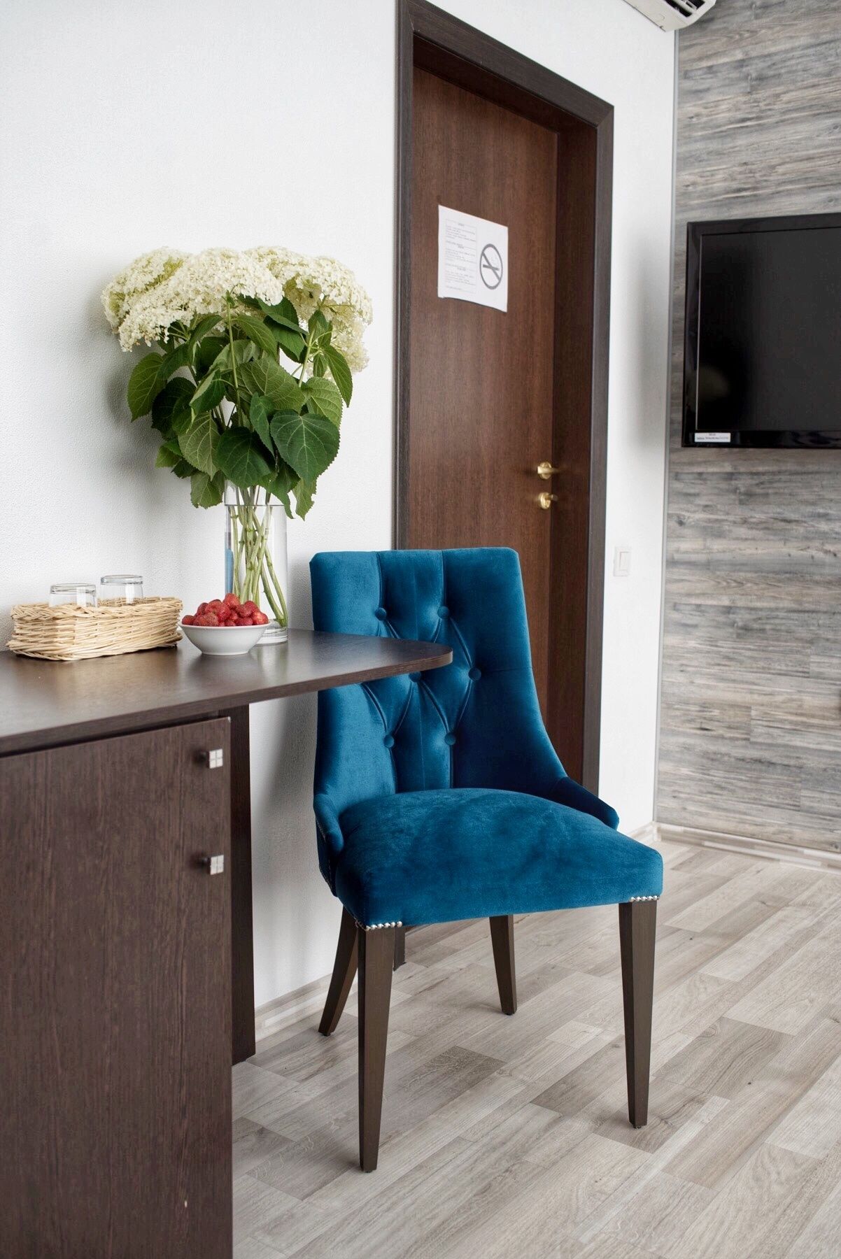 A room with a dark wooden desk, a blue velvet chair, a white vase with white flowers, and a closed door with a sign.
