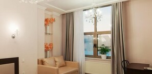 A living room with a beige sofa, a chandelier, a window with curtains, and a potted plant on the windowsill. - Hermes Hotel (Odesa)