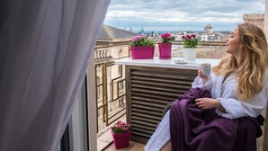 Eine Person sitzt auf einem Balkon mit Blumentöpfen und einer Tasse und blickt auf eine Stadtansicht.