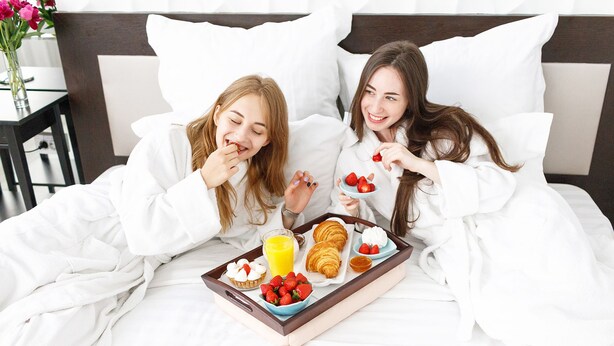 Zwei Personen in einem Schlafzimmer mit einem Tablett mit Frühstücksangeboten, darunter Croissants, Erdbeeren und ein Glas Orangensaft.