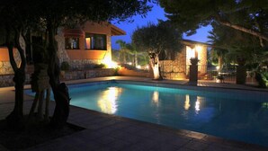 A swimming pool with a stone wall and a building with warm lighting in the background. - Borgo dei Vigneti (Agrigento)