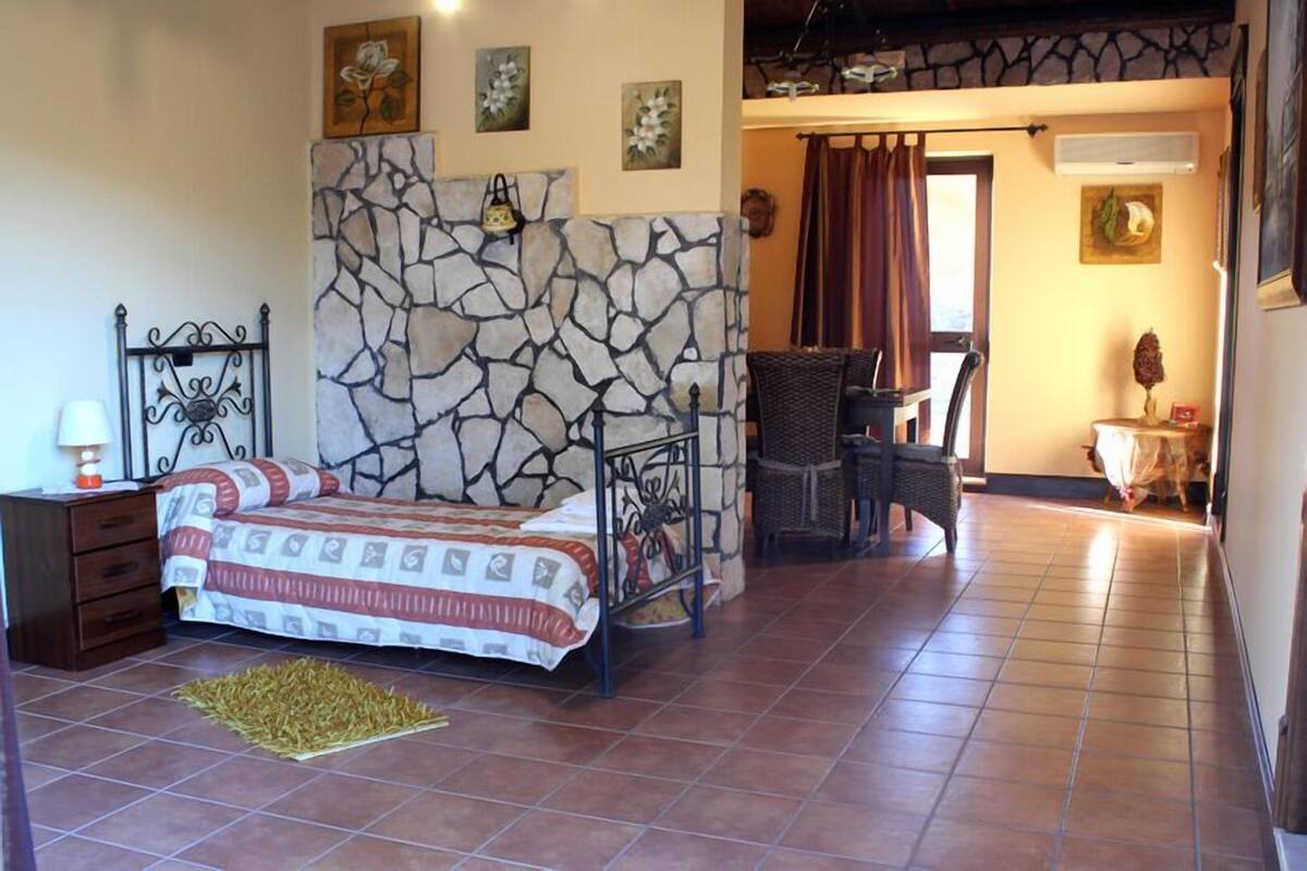 A bedroom with a stone wall, a bed with a patterned bedspread, a nightstand, a chair, and a small table with a lamp.