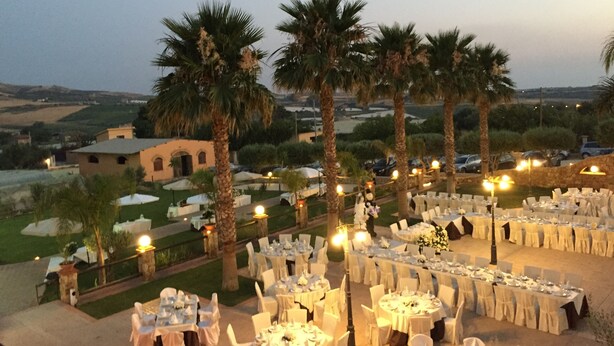An outdoor dining area with multiple round tables set up for guests, surrounded by palm trees and a view of the countryside. - Borgo dei Vigneti (Agrigento)