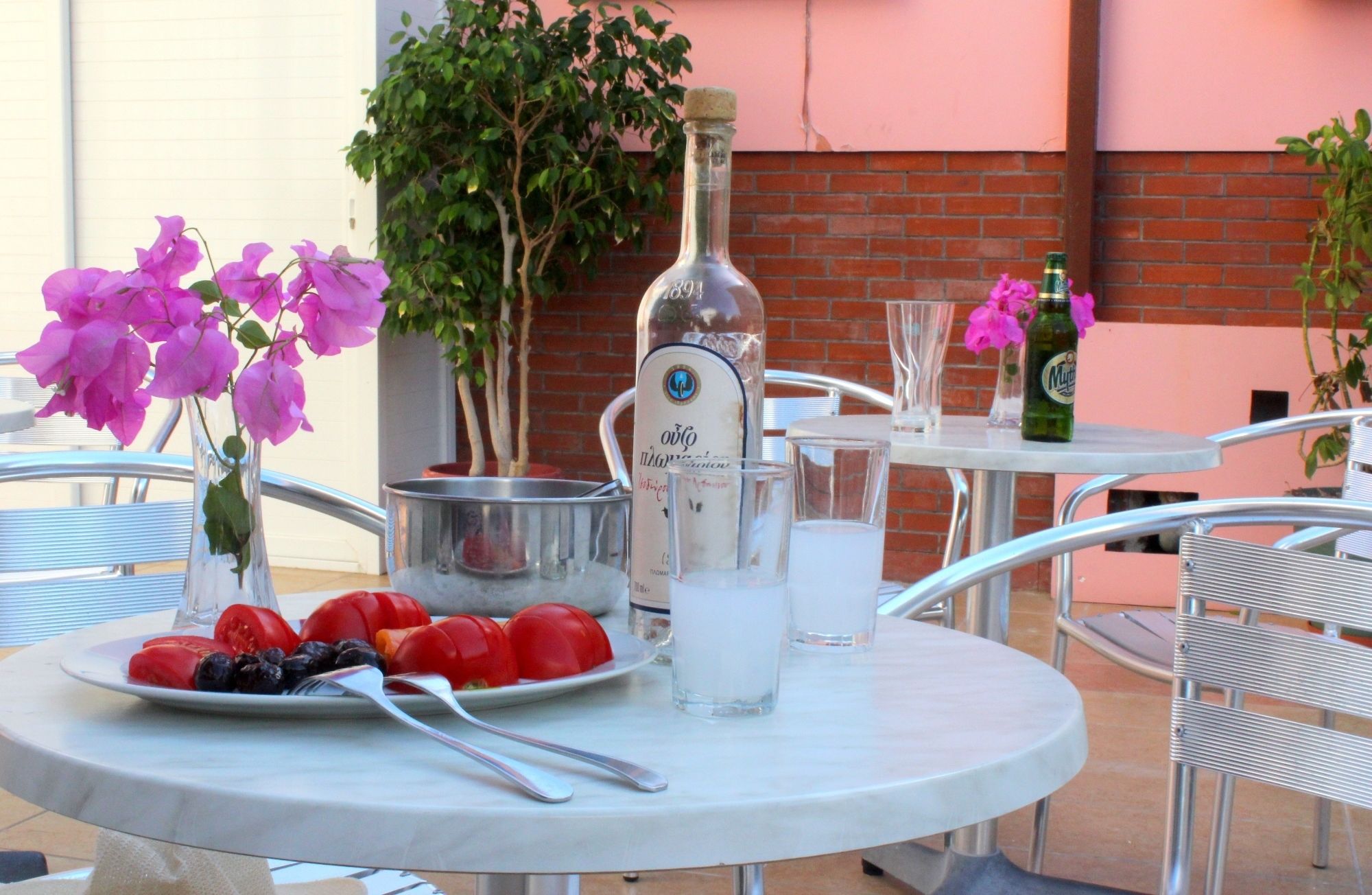 Uma mesa redonda ao ar livre com uma garrafa de vinho, um balde de metal, uma travessa de frutas e um copo de água.