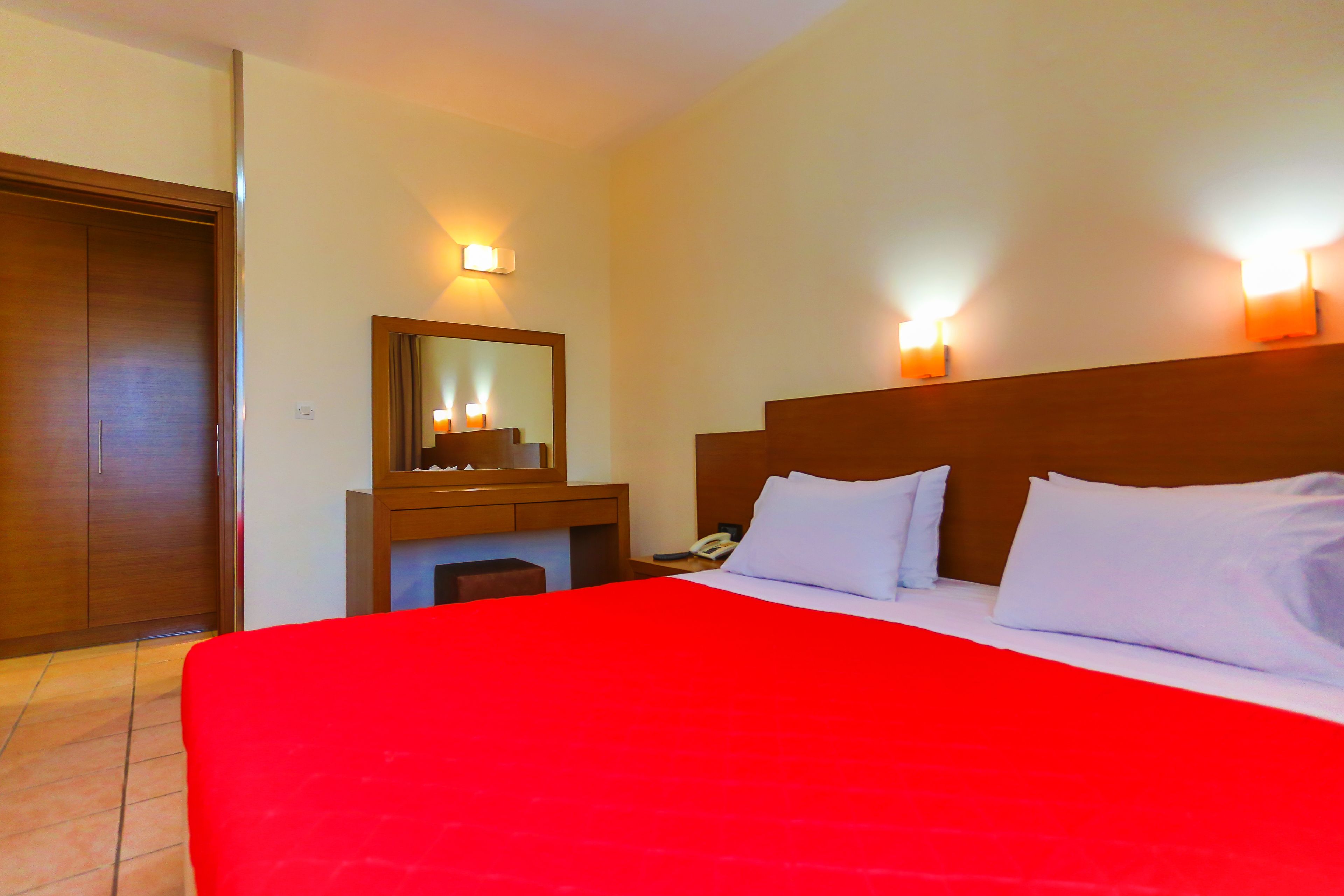 A hotel room with a large bed, a wooden headboard, a nightstand with a phone, a mirror, and wall-mounted lights.