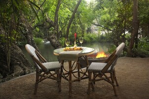 Un área de comedor al aire libre con sillas de mimbre y una mesa, rodeada de exuberante vegetación y una masa de agua.