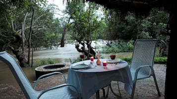 An outdoor dining area with a table set for two, surrounded by chairs, with a view of a river and dense foliage.