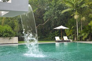 Una piscina con cascada, rodeada de exuberante vegetación y un área de descanso con sillas y una sombrilla.