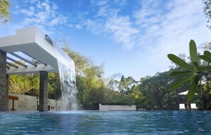 A swimming pool with a waterfall feature, surrounded by greenery and a clear sky. - Amanvana Spa Resort - Coorg (Somvarpet)