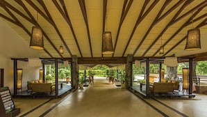 A spacious lobby with a high ceiling, wooden beams, and hanging lamps. There are seating areas with tables and chairs, and a view of the outdoors through large windows. - The Windflower Resorts & Spa (Somvarpet)