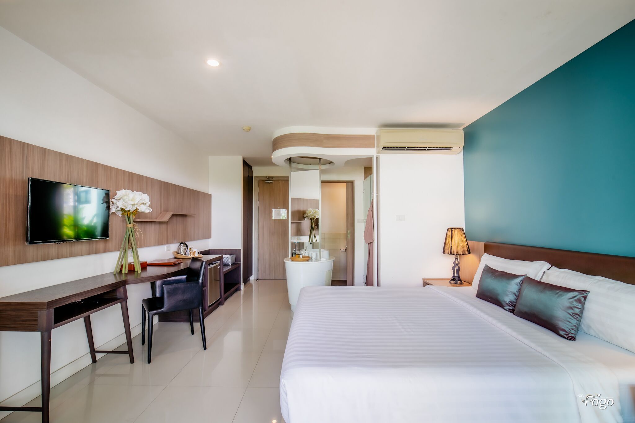 A modern hotel room with a large bed, a desk with a chair, a TV, and a bathroom visible through an open door.