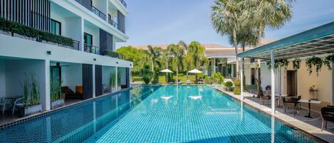 Una piscina con solárium de baldosas, rodeada por un edificio moderno con balcones y mobiliario exterior.