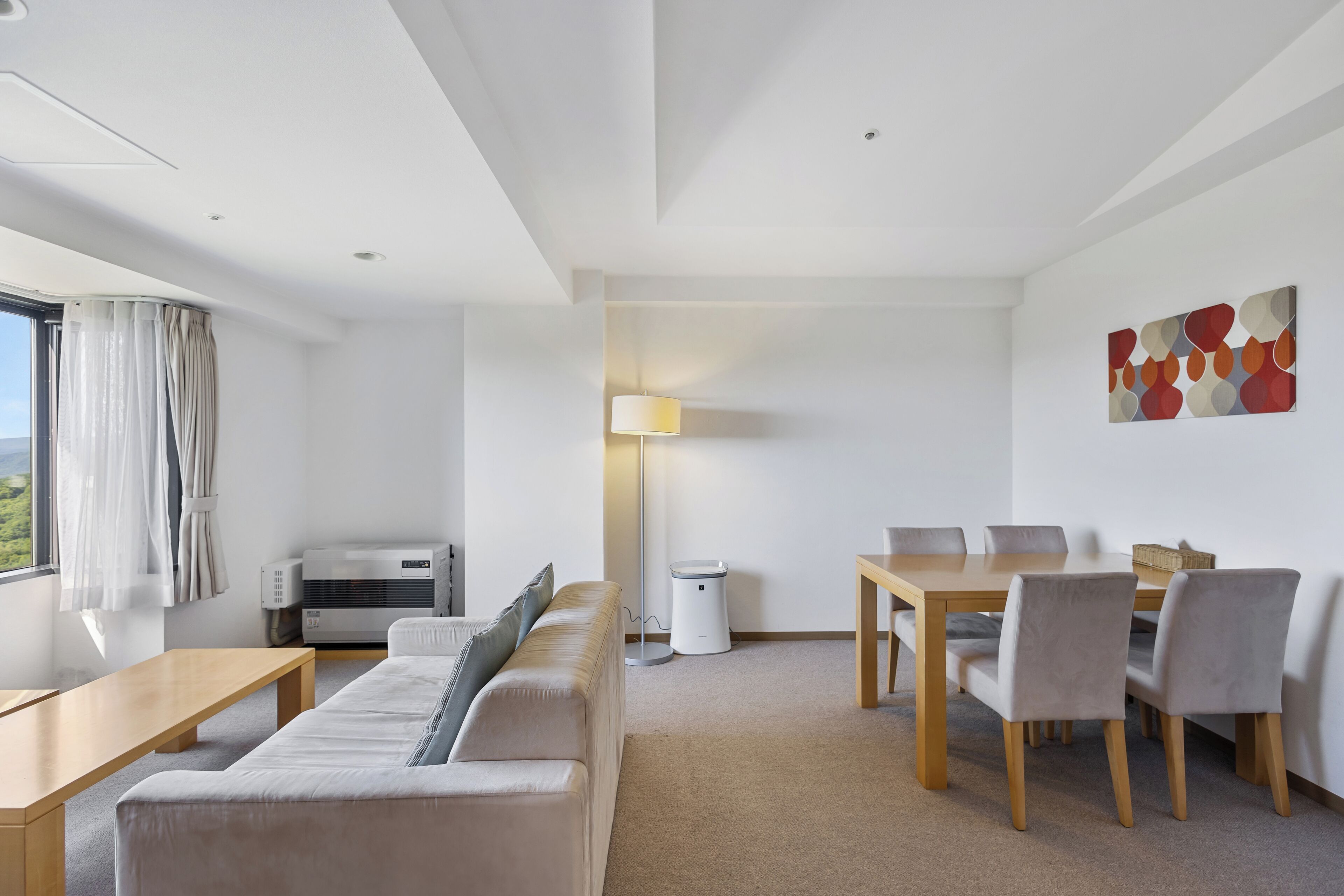 Una habitación de hotel moderna con sofá, una mesa de comedor con sillas, una lámpara y un gran ventanal con cortinas.