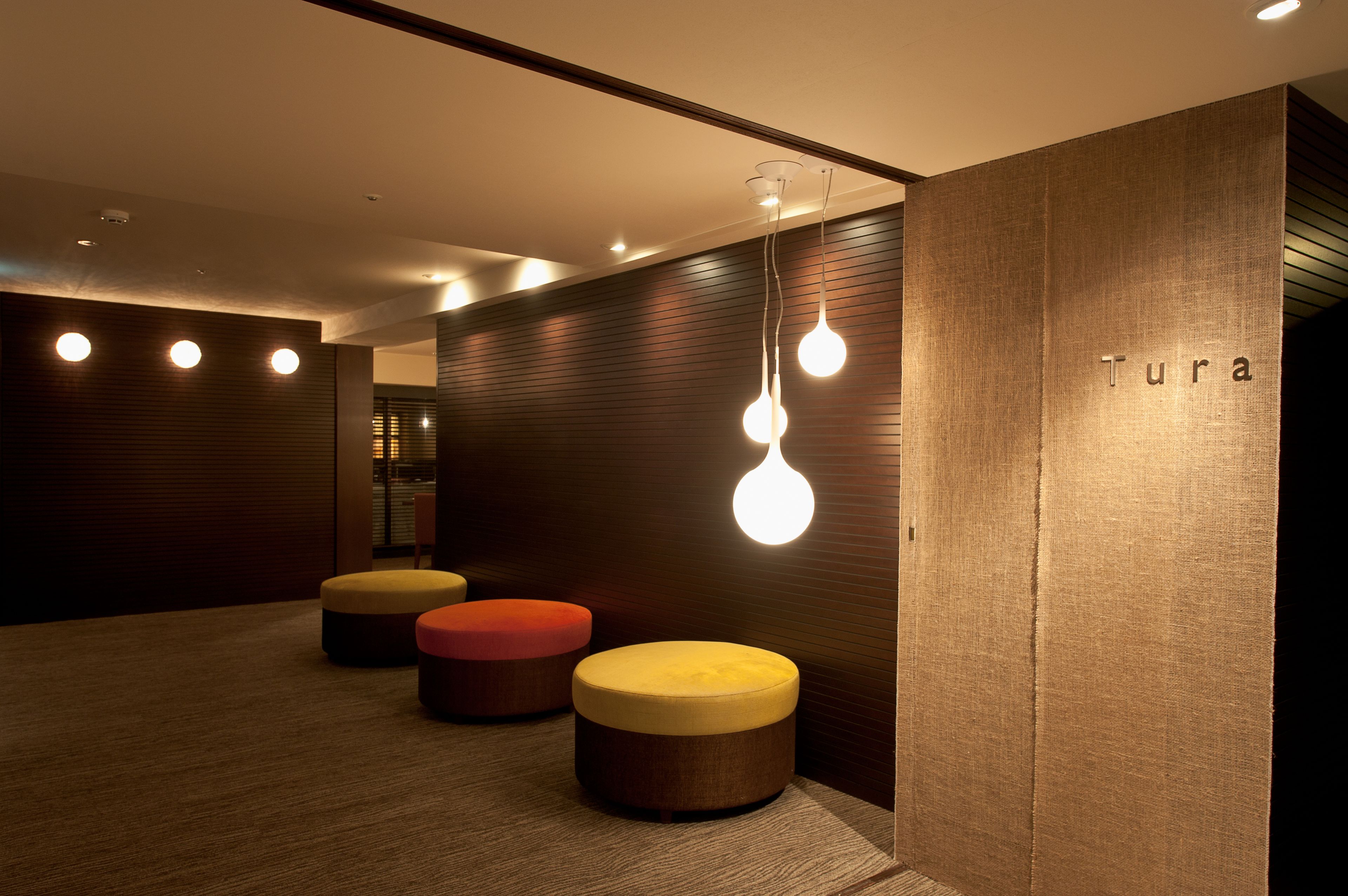 A modern interior with wooden panel walls, a reception desk labeled "Tura," and three round ottomans in red, yellow, and brown.