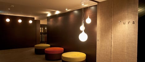 A modern interior with wooden panel walls, a reception desk labeled "Tura," and three round ottomans in red, yellow, and brown.