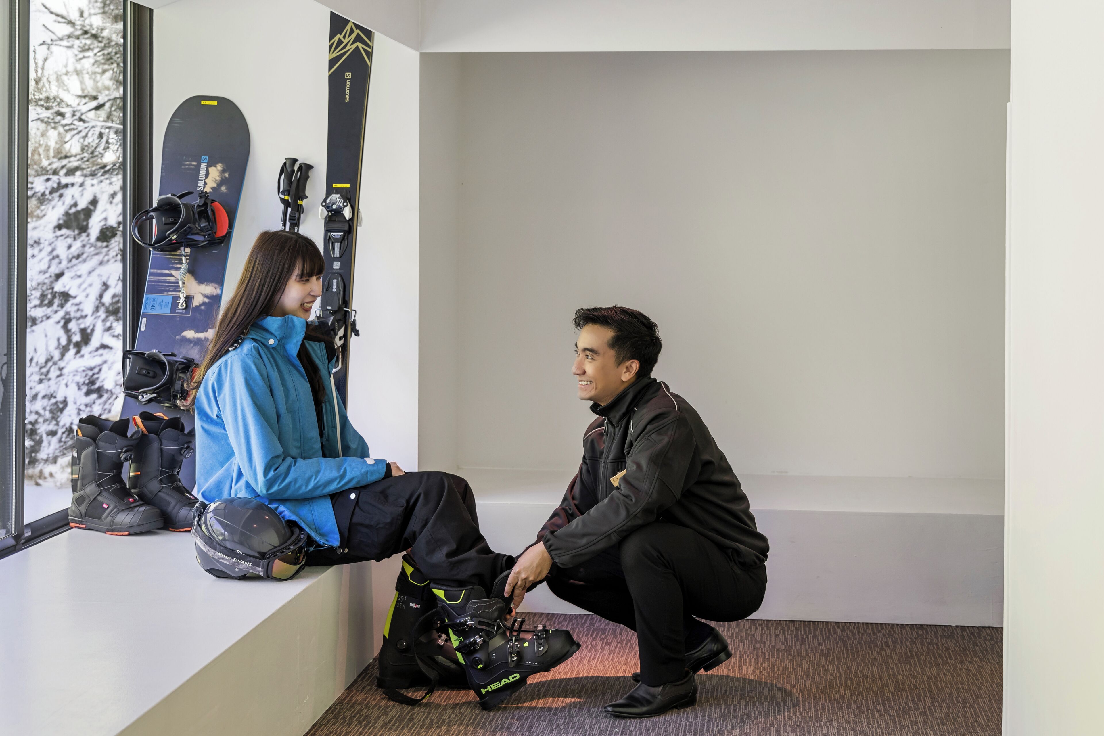A room with a window overlooking a snowy landscape, a pair of skis leaning against the wall, and a person sitting on a ledge.