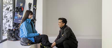 A room with a window overlooking a snowy landscape, a pair of skis leaning against the wall, and a person sitting on a ledge.