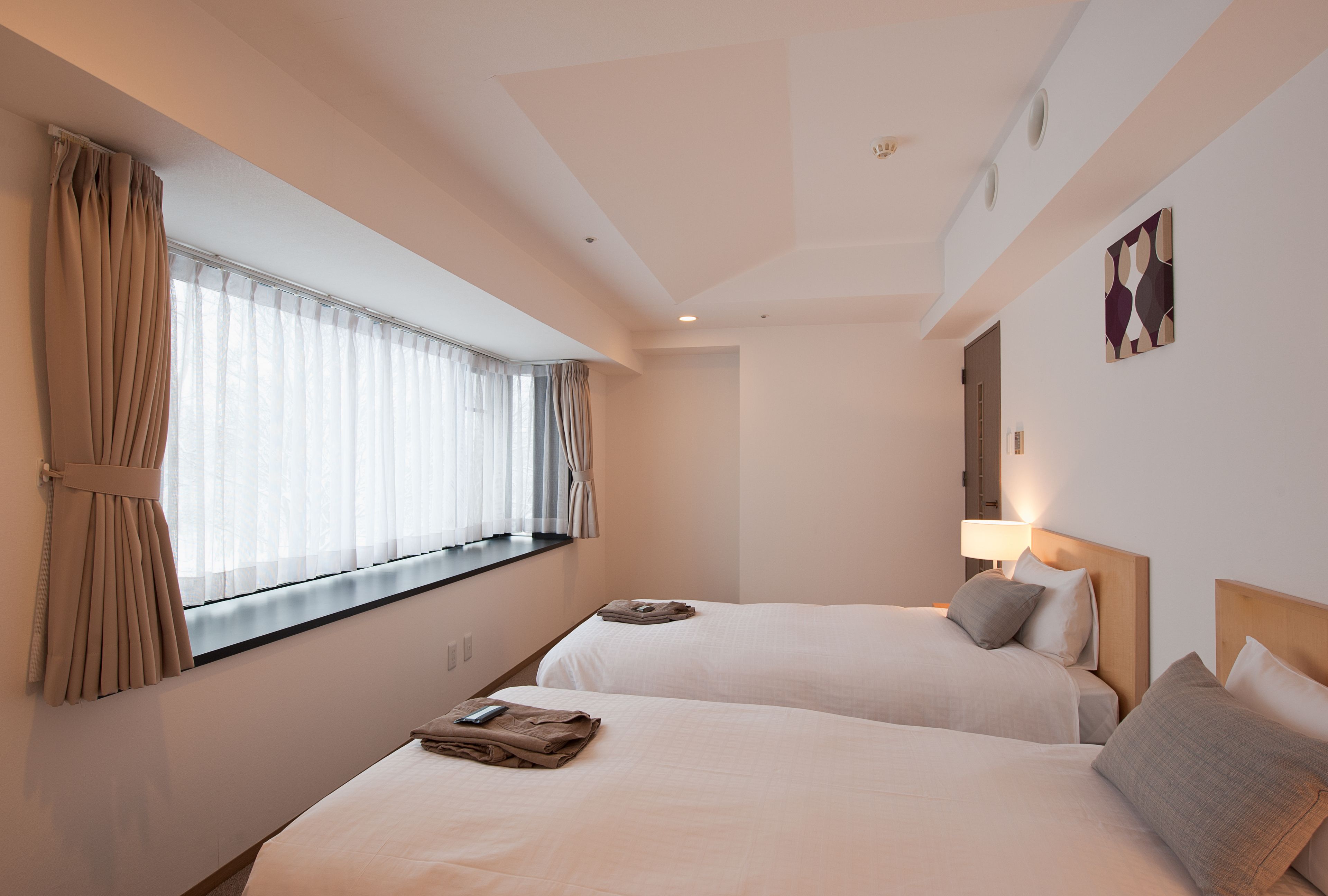 A hotel room with two beds, a window with curtains, a wall-mounted lamp, and a painting.