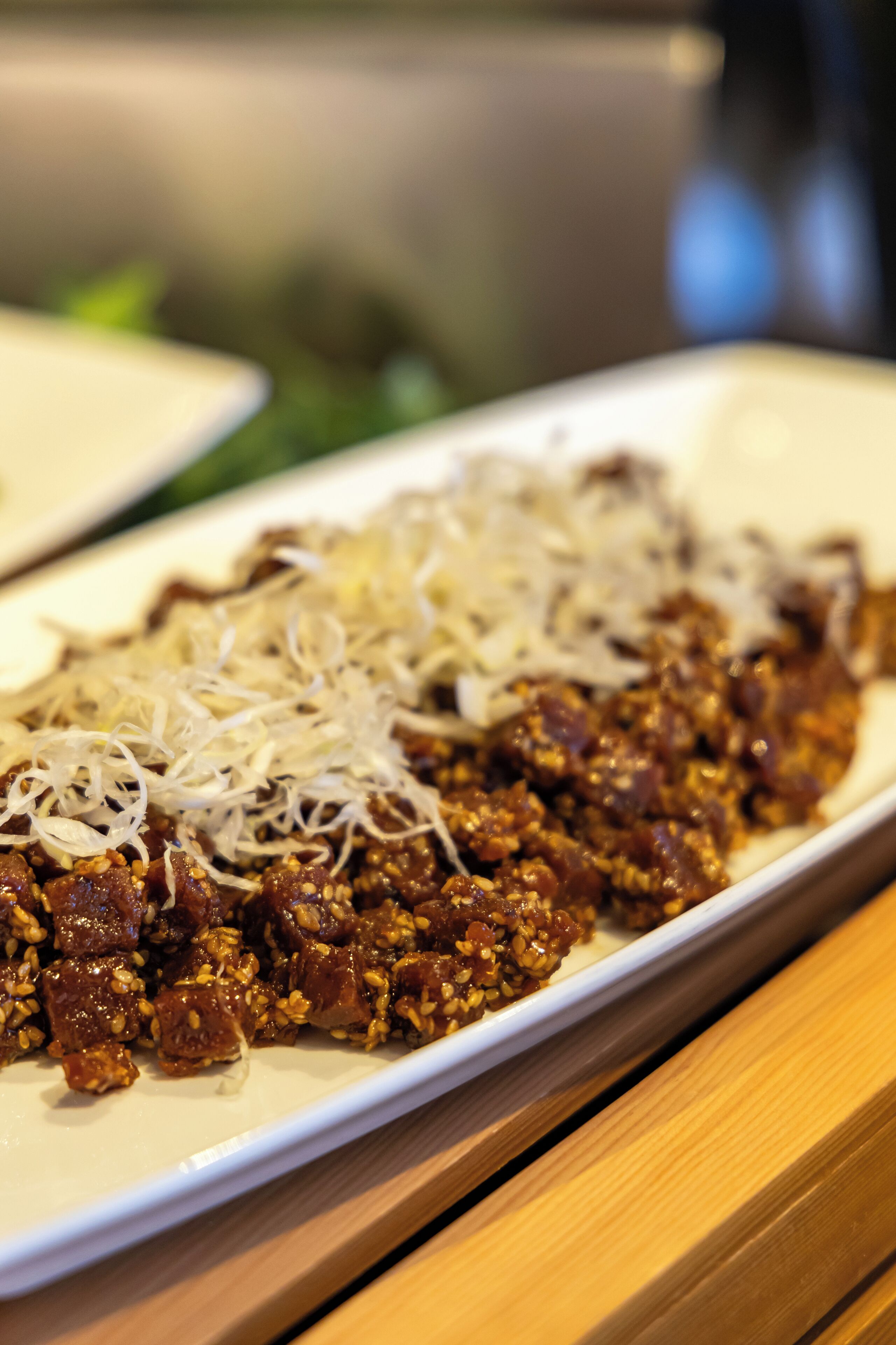 A rectangular plate with a dish that includes shredded white vegetables and a dark, grainy topping, possibly meat, on a wooden table.