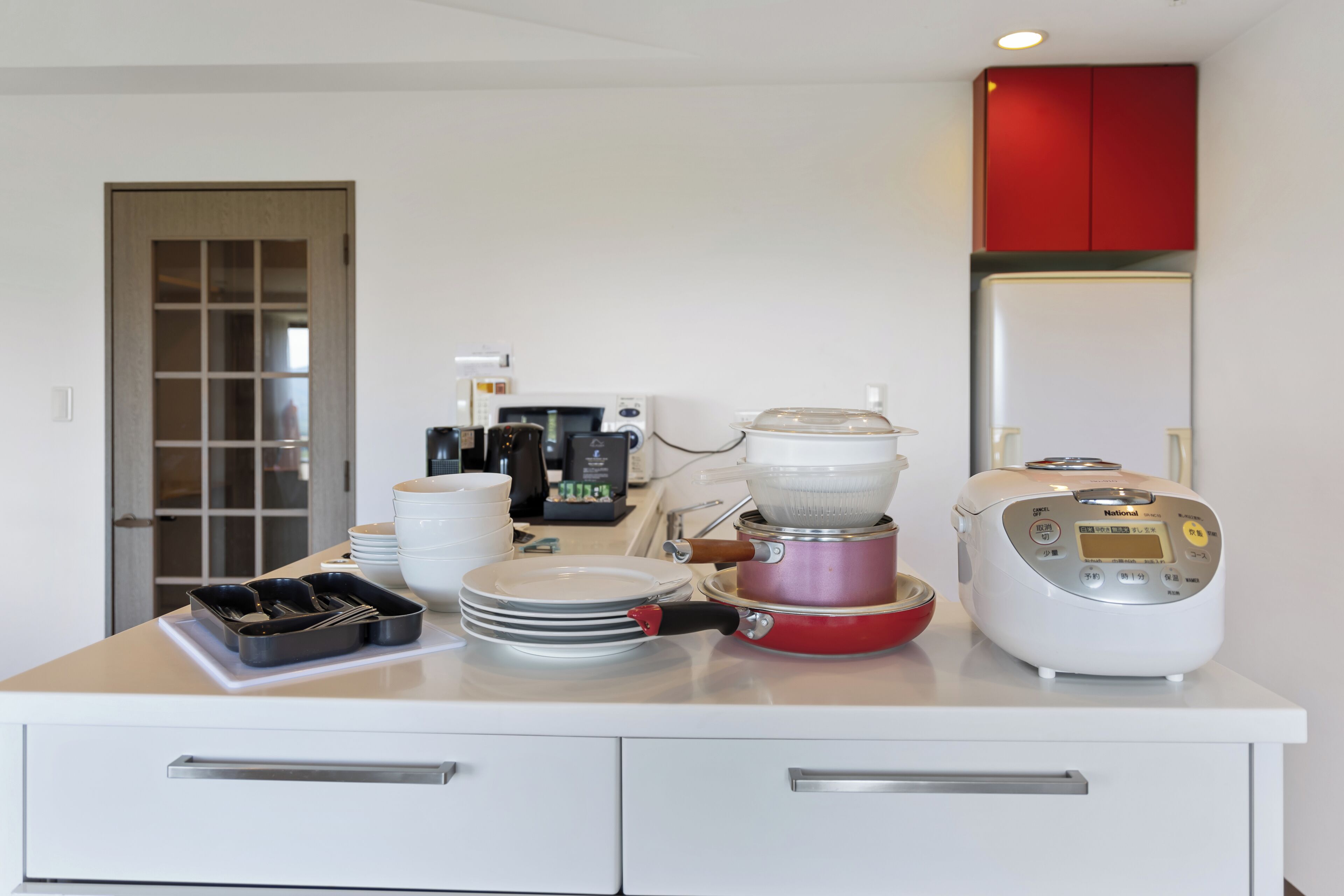 A kitchen with a microwave, coffee maker, rice cooker, and a red cabinet above the counter.