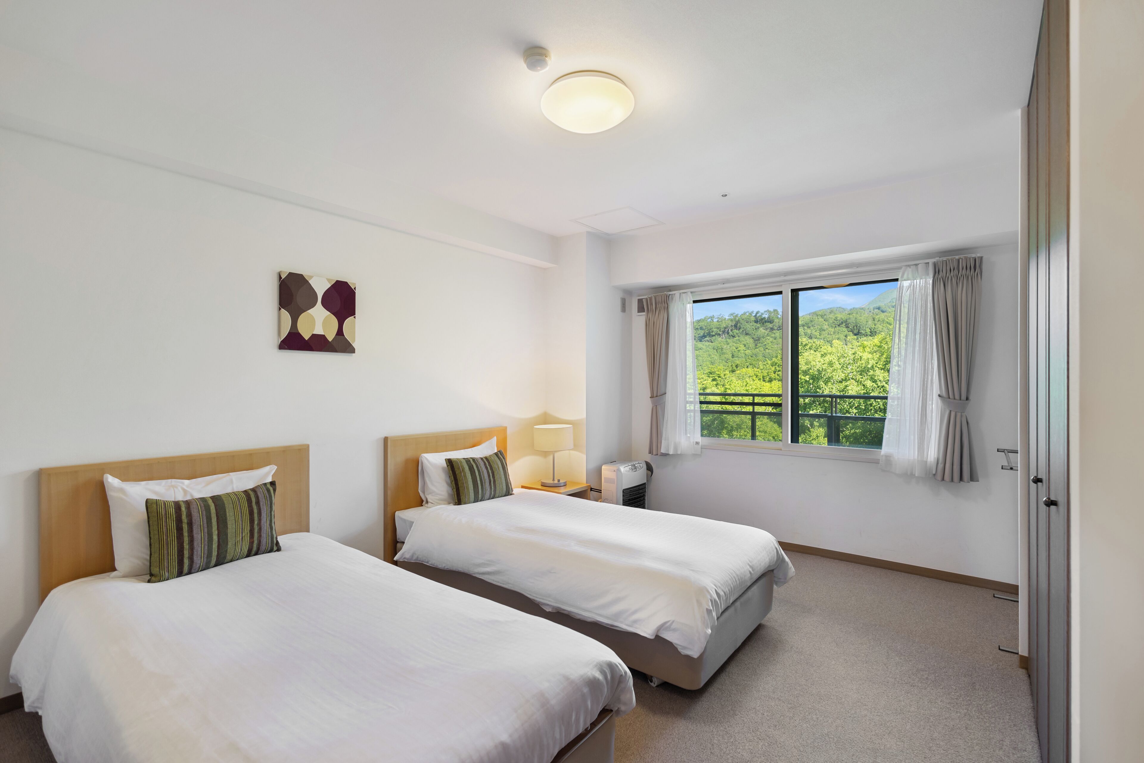A hotel room with two beds, a window with a view of greenery, a ceiling light, and a wall-mounted artwork.