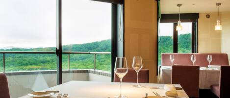 A dining area with a table set for two, two wine glasses, and a view of a lush green landscape through large windows.