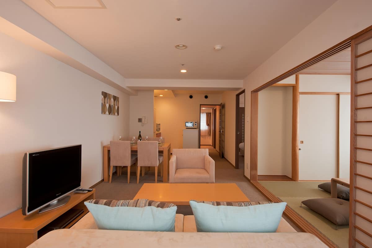 Ein modernes Hotelzimmer mit Flachbildfernseher, einer Couch mit Kissen, einem Essbereich mit Tisch und Stühlen sowie einer traditionellen Schiebetür.