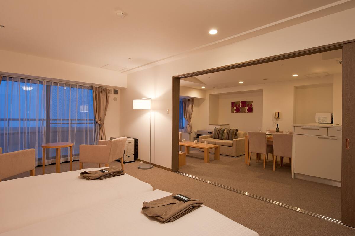 Ein Hotelzimmer mit einem großen Bett, einem Schreibtisch, einem Sessel, einer Couch und einem Esstisch. An einer Wand befindet sich ein Spiegel und durch die Vorhänge ist der Balkon zu sehen.