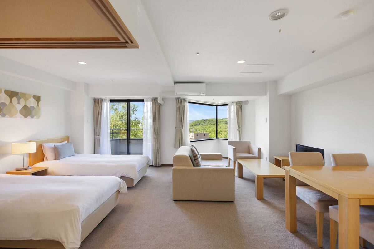 Ein Hotelzimmer mit zwei Betten, einem Sofa, einem Esstisch und großen Fenstern mit Blick auf Grünflächen.