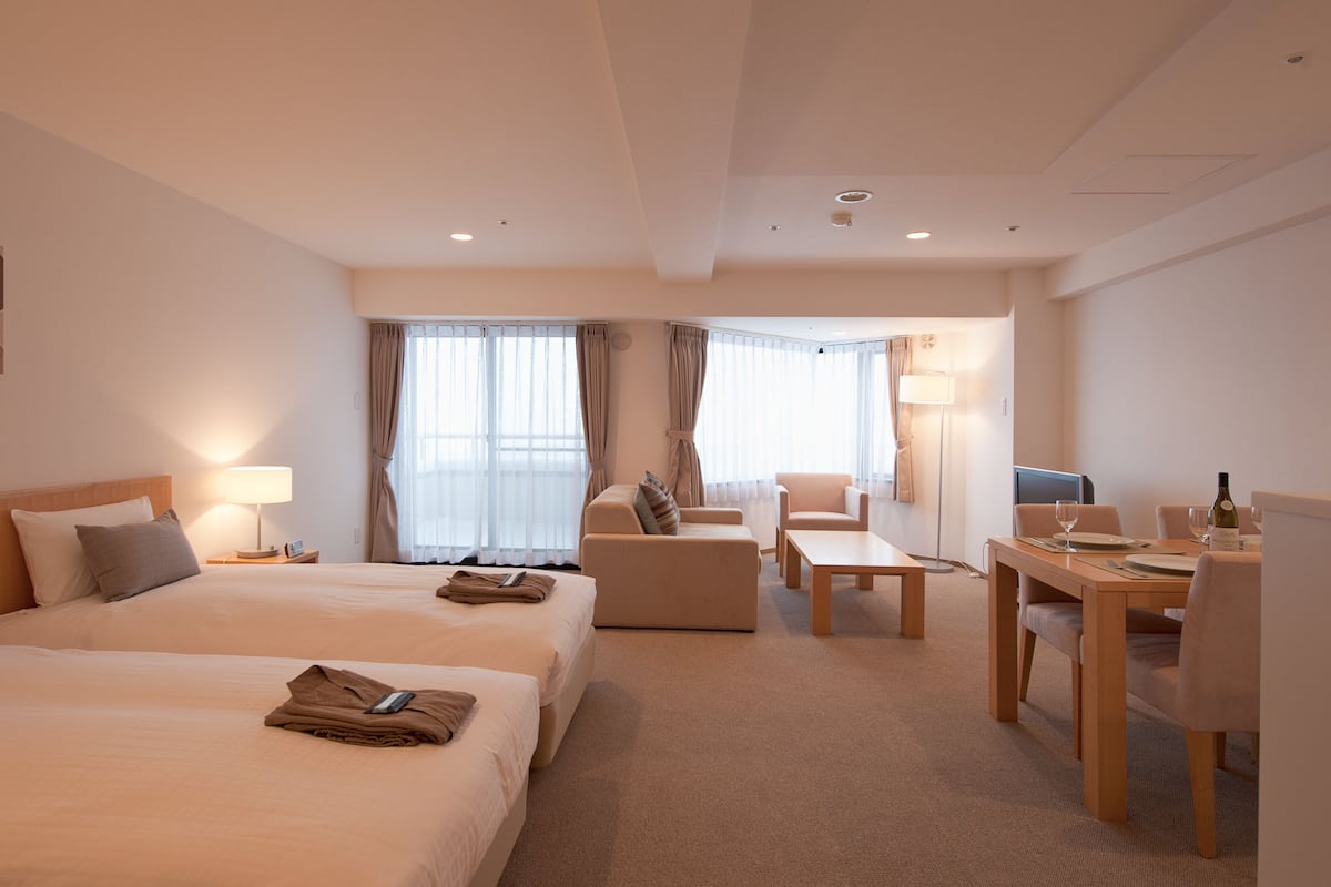Ein Hotelzimmer mit zwei Betten, einem Schlafsofa, einem Esstisch mit Stühlen, einer Lampe und einem Fernseher.