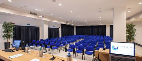 A conference room with rows of blue chairs, a wooden table with a laptop, a projector, and a water dispenser.
