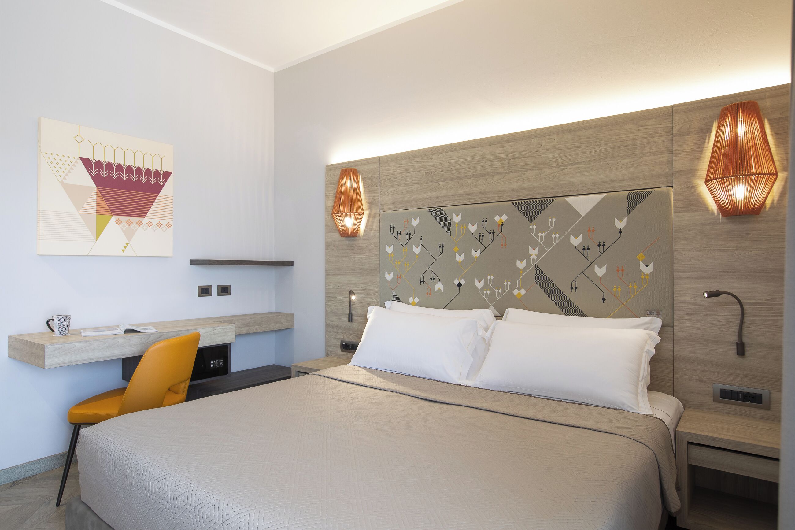 A modern hotel room with a large bed, a desk with a chair, and decorative wall art.
