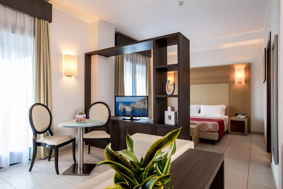 A hotel room with a desk, chair, TV, bed, bedside table, and a plant.