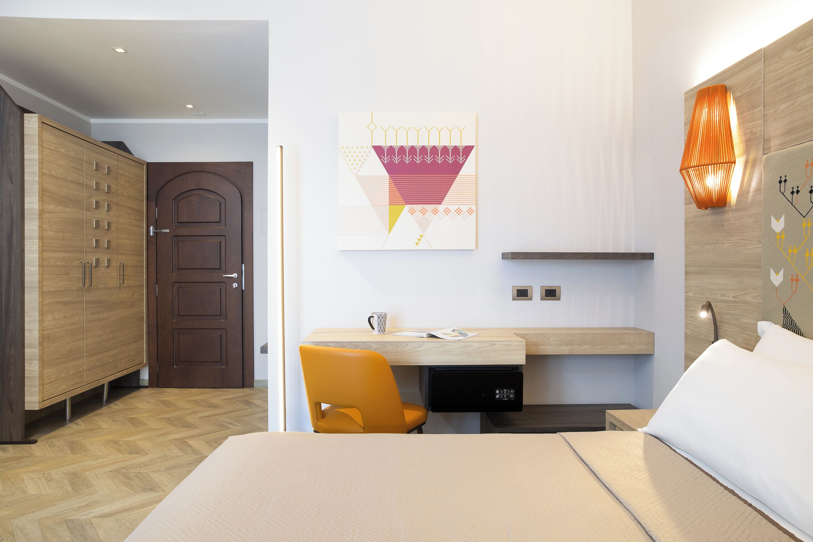 A modern hotel room with a wooden wardrobe, a desk with a chair, a bed, and a wall-mounted artwork.