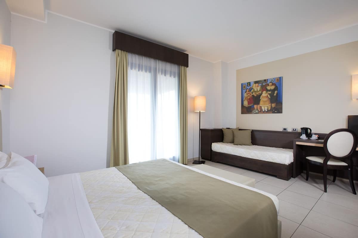 A hotel room with a bed, a desk, a chair, a painting on the wall, and a window with curtains.