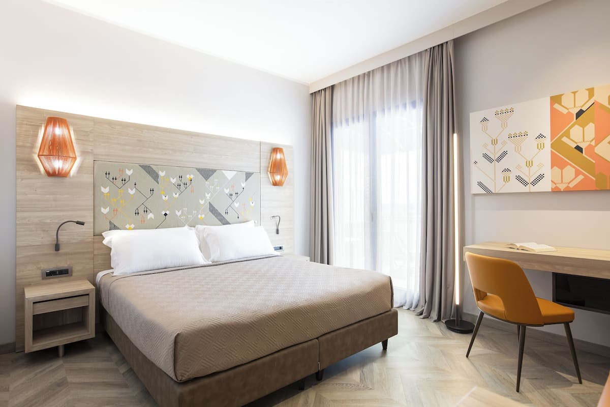 A modern hotel room with a large bed, a desk, and a chair. There are two wall-mounted lamps, a painting, and a window with curtains.