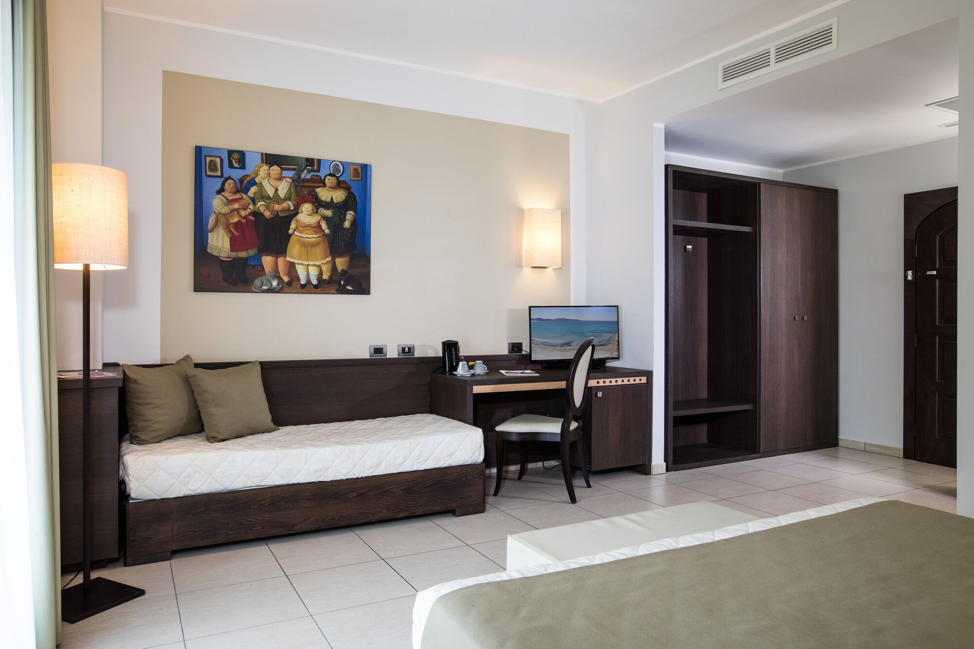 Une chambre d’hôtel avec un lit, un bureau avec un ordinateur, une chaise, une armoire et un tableau au mur.