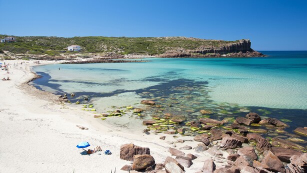 A beach with clear turquoise water, rocky shoreline, and a few buildings in the background. - Lu' Hotel Carbonia (Carbonia)