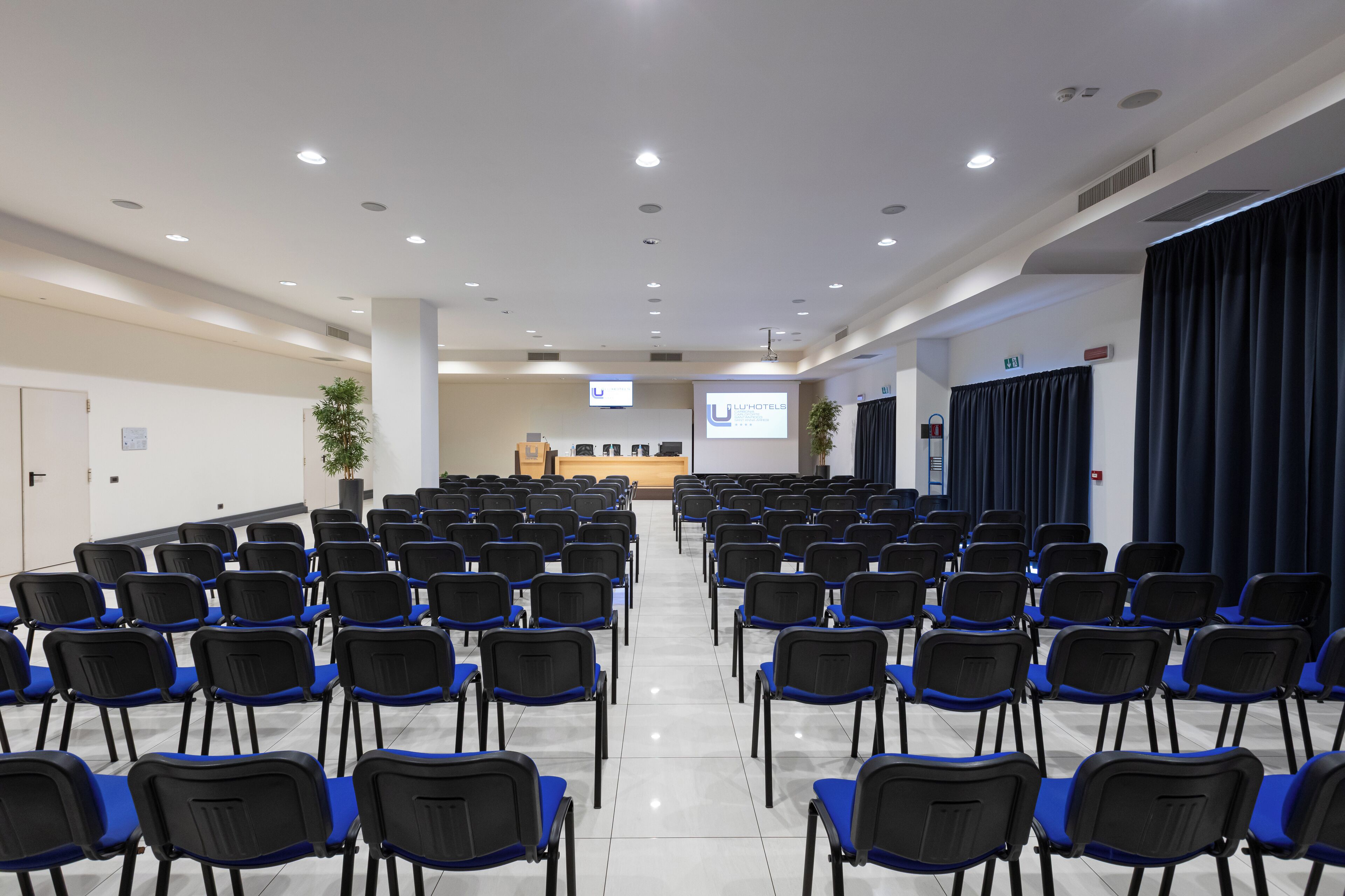 Een vergaderzaal met rijen stoelen, een spreekgestoelte en een projectiescherm.