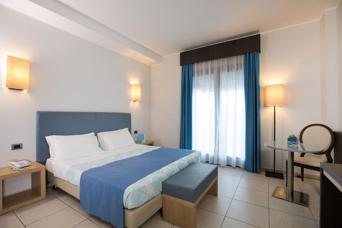 A hotel room with a bed, bedside tables, a desk with a chair, a lamp, and a window with curtains.