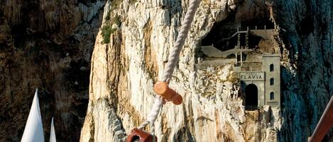 A boat with ropes and a sailboat in the background, anchored near a rocky cliff with a small building labeled "Porto Flava".