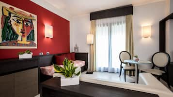 A modern hotel room with a red accent wall, a large abstract painting, a sofa, a dining table with chairs, and a window with curtains.
