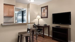 A compact living space with a kitchenette, a dining area with bar stools, a desk with a lamp, and a mounted TV. - Eagle's Den Suites Carrizo Springs a Travelodge by Wyndham (Carrizo Springs)
