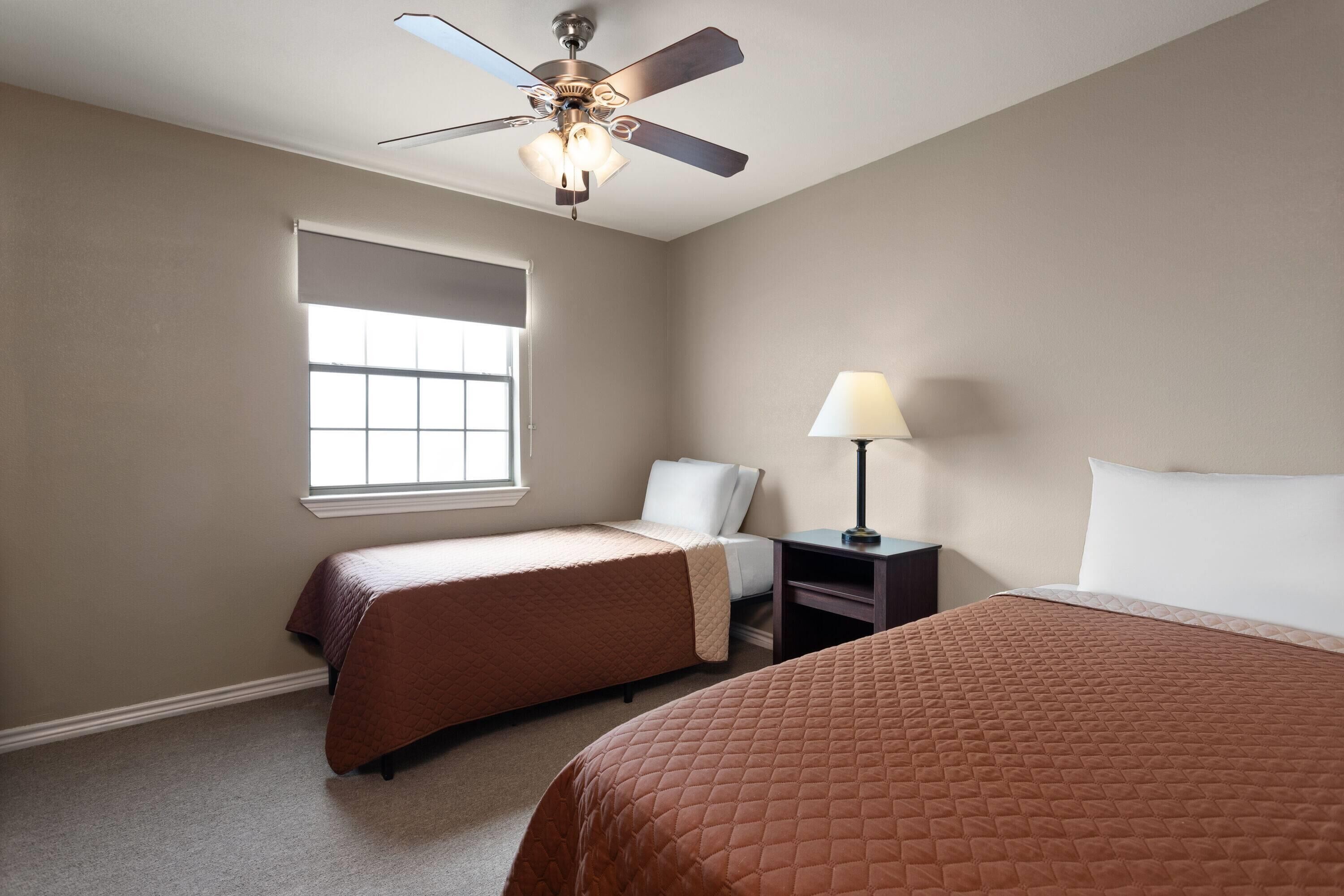 Quarto de hotel com duas camas, um ventilador de teto, uma janela com persianas, uma mesa de cabeceira com abajur e uma mesinha de apoio.