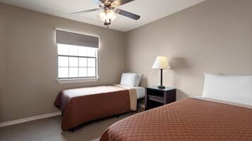 Quarto de hotel com duas camas, um ventilador de teto, uma janela com persianas, uma mesa de cabeceira com abajur e uma mesinha de apoio.