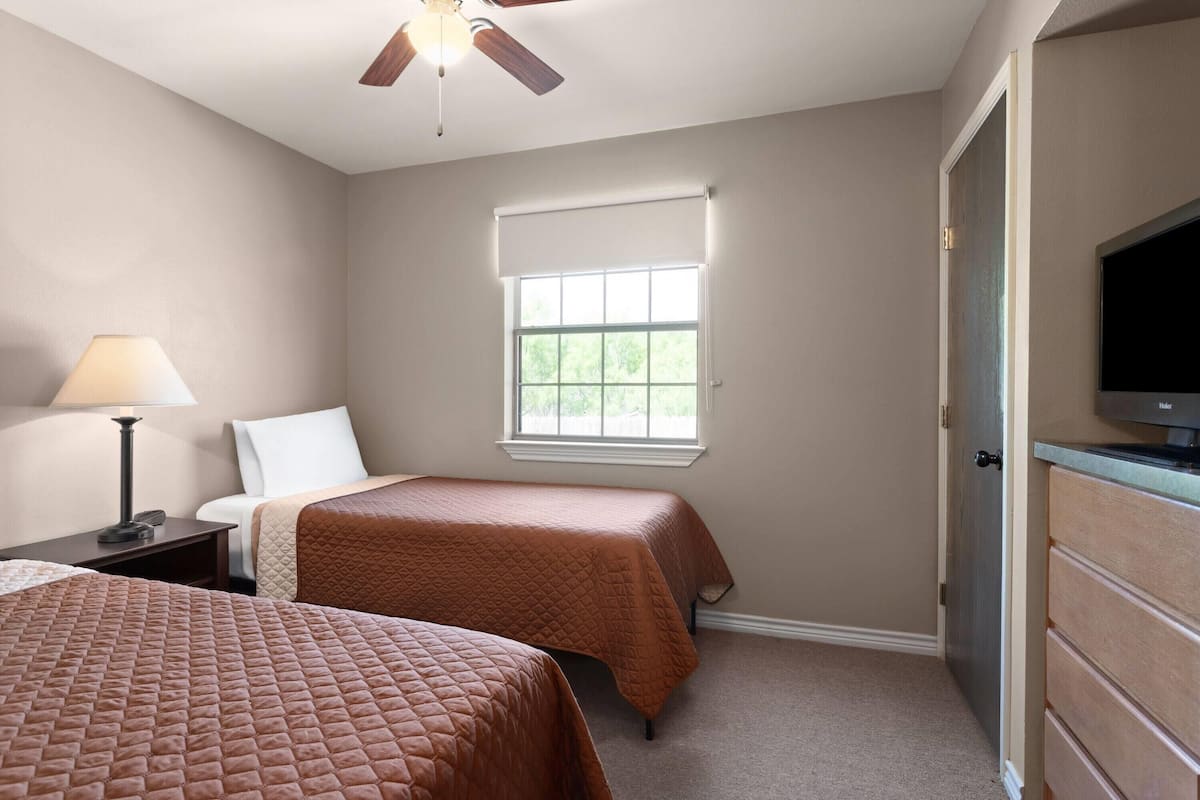 A hotel room with two beds, a ceiling fan, a window with blinds, a television, and a desk with a lamp.