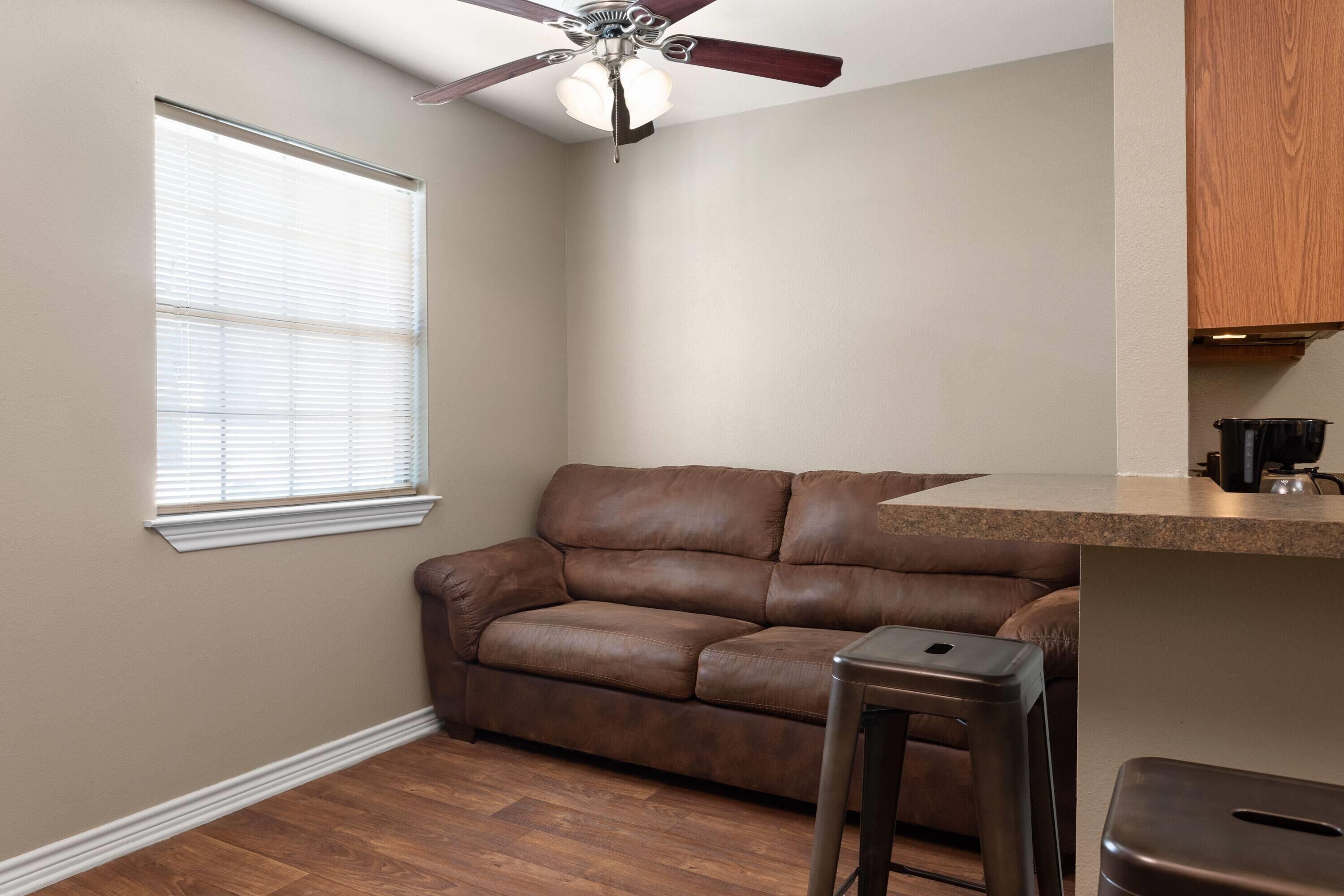 A room with a brown leather sofa, a ceiling fan, a window with blinds, a kitchen counter, and a stainless steel trash can.