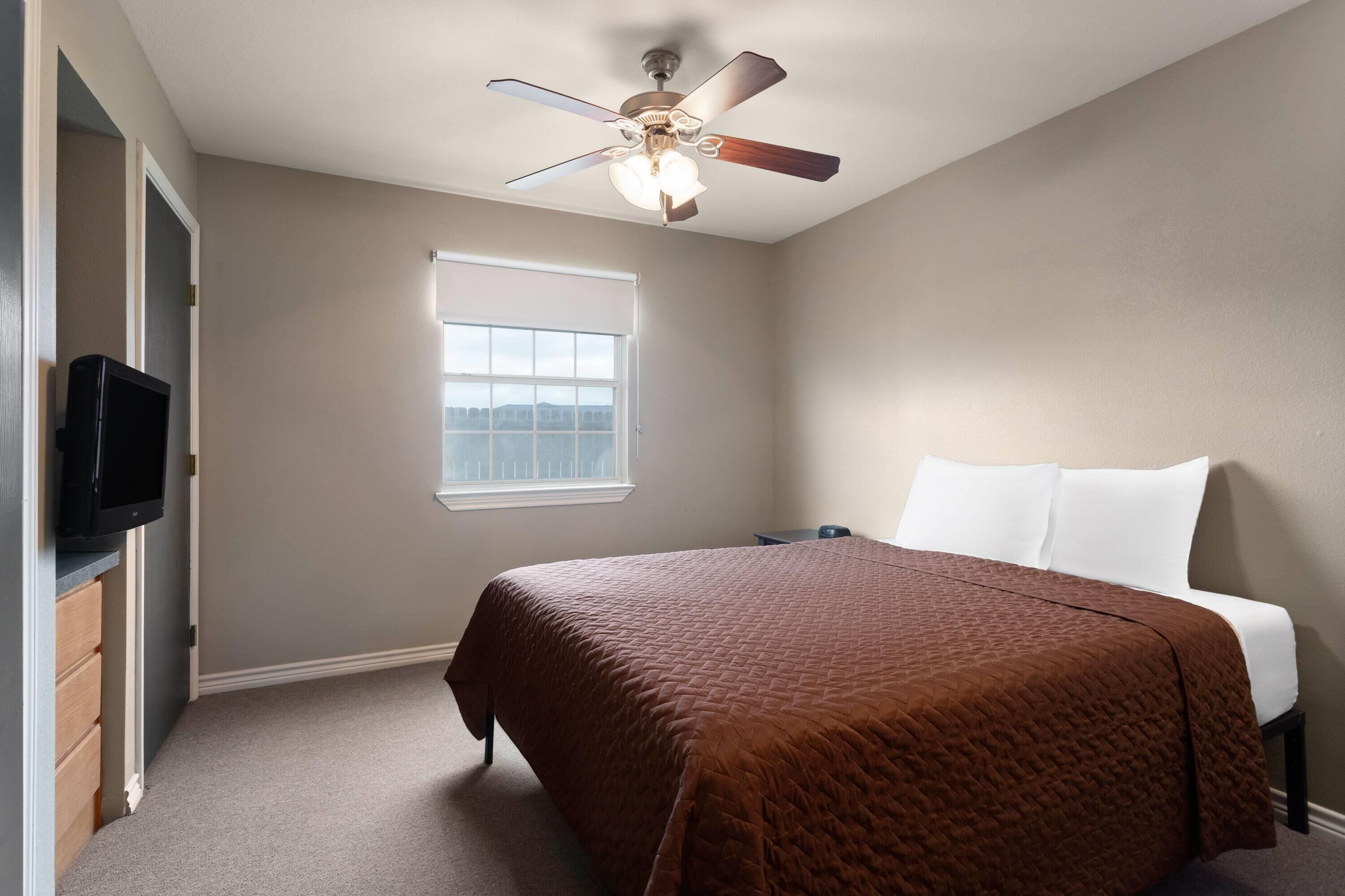 Un dormitorio con cama, ventilador de techo, ventana y televisor sobre un aparador.